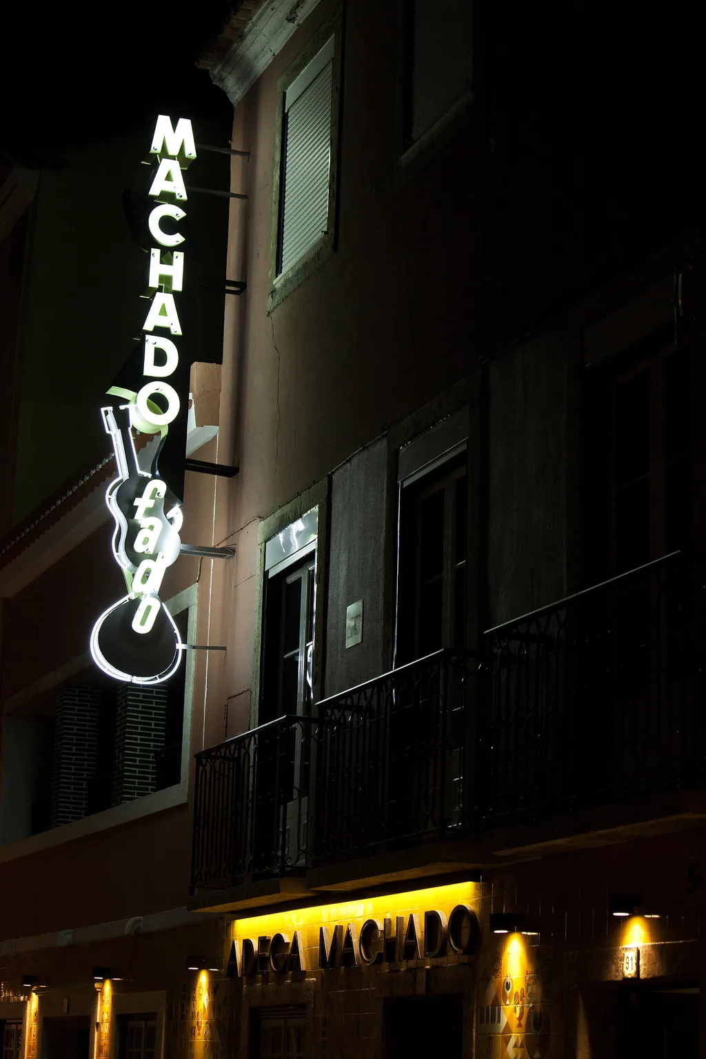 Letreiro de néon aceso sobre a fachada amarela iluminada da Adega Machado