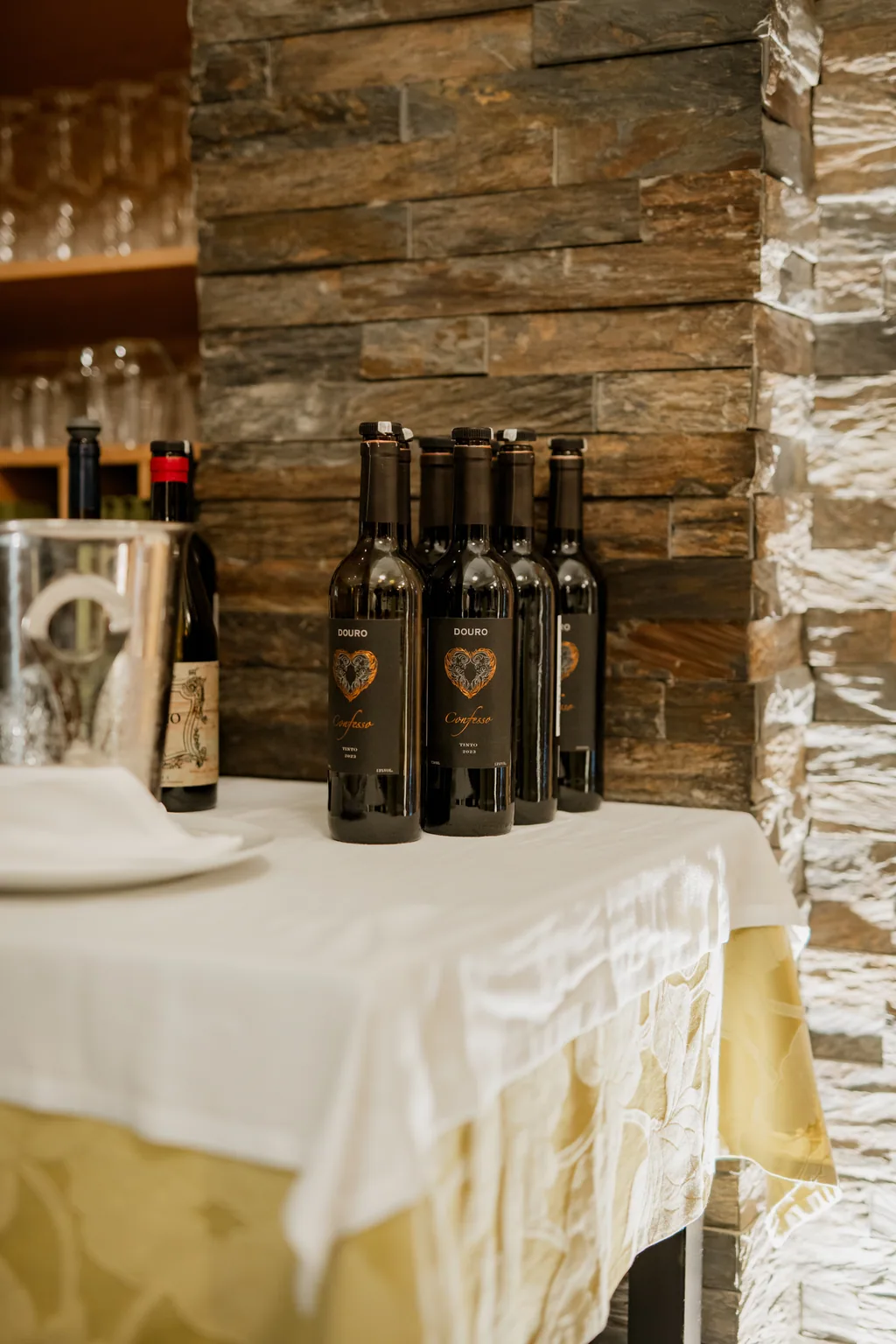 Fila de garrafas de vinho tinto sobre a toalha branca, junto à parede de pedra