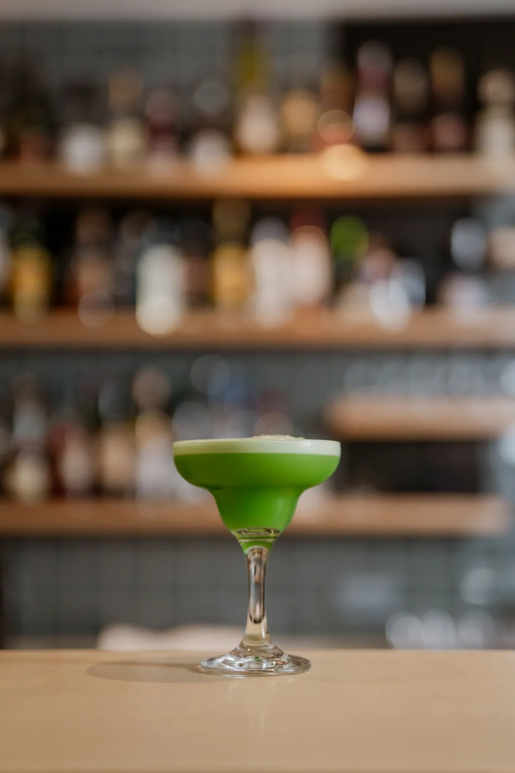 Cocktail verde em taça coupe sobre o balcão, com as garrafas desfocadas atrás