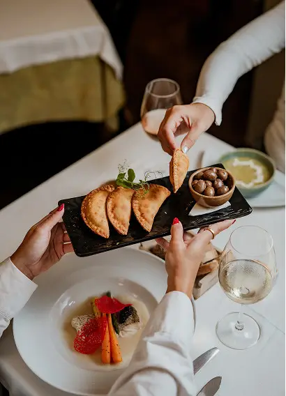 Duas pessoas jantam à mesa, uma passando um prato retangular de empanadas e azeitonas para a outra. Um copo de vinho branco, legumes gourmet e peixe do menu do chefe também estão na mesa elegantemente posta.