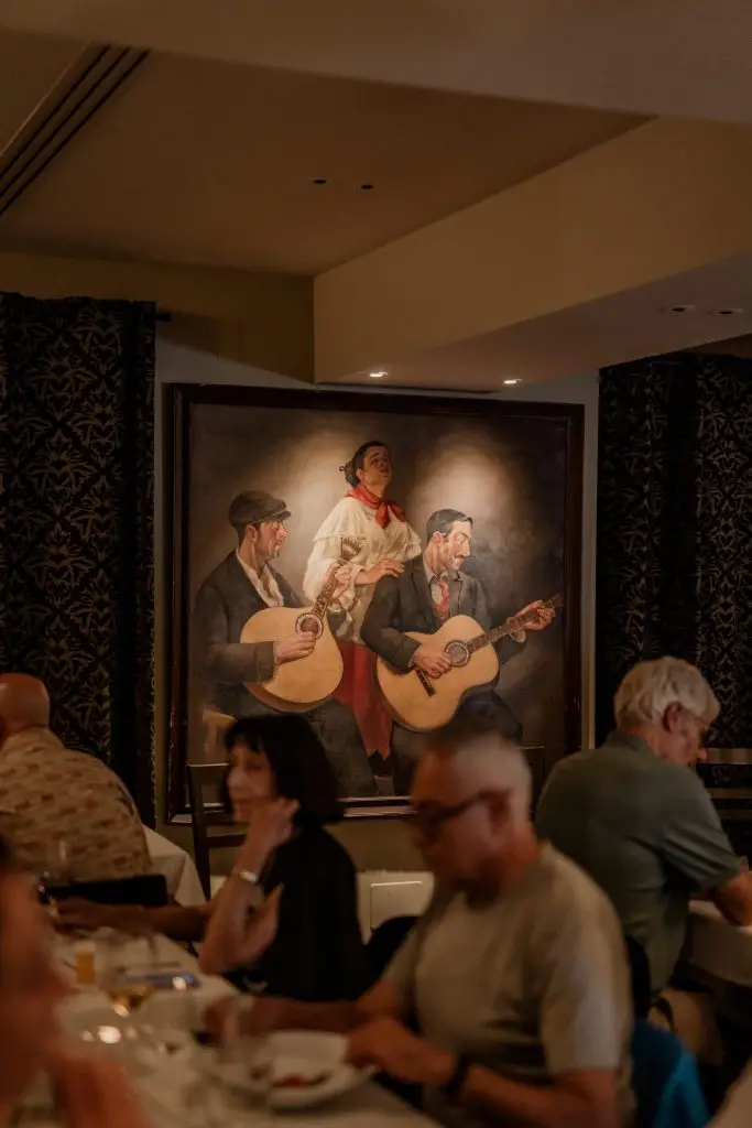 Clientes a jantar na sala da Adega Machado com quadro de músicos a tocar guitarra portuguesa pendurado ao fundo, Lisboa